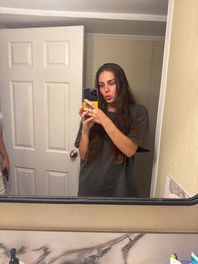 A young woman with long, dark hair and an olive complexion is taking a mirror selfie in a bathroom. She is wearing a dark gray t-shirt and a delicate gold bracelet on her left wrist. Her lips are pursed, and she is holding a smartphone with a yellow case in her right hand to capture the reflection. The background shows a white paneled door and a textured beige wall. The bottom of the image displays a portion of a marble-patterned countertop with various toiletries, including a pump bottle, a pink object, and a tube of toothpaste. The overall scene suggests a casual moment in a domestic setting. The specific location of Ocean City, United States of America is not discernible from the provided image.