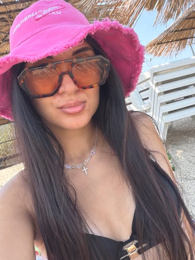 In an outdoor, beachfront setting, a person is taking a selfie. The subject is wearing a bright pink bucket hat with frayed edges, large, tortoiseshell-patterned sunglasses with amber lenses, and a black bikini top with gold hardware. A delicate necklace with clear beads and a small silver cross pendant adorns their neck. Their long, dark hair is visible, framing their face and falling over their shoulders. In the background, beach chairs and umbrellas are visible, suggesting a sunny day at the beach. The location is identified as Pavlo-Ochakovo, Russia.