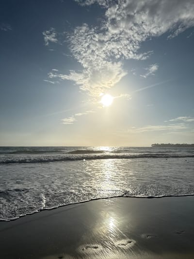 The image shows a beach scene in Cabarete, Dominican Republic. The sun is high in the sky, partially obscured by clouds. The ocean is visible, with waves breaking on the shoreline. The sand is wet and reflective, with footprints visible near the edge of the water.