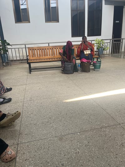 This image, taken in Picha Ya Ndege, Tanzania, depicts a waiting area, possibly in a public building like a clinic or travel hub. The scene is illuminated by a strong, diagonal beam of natural sunlight cutting across the speckled terrazzo floor.

In the mid-ground, two individuals are seated on a long, light wooden bench. On the left, an individual, appearing to be an older man, wears a vibrant red, black, and yellow patterned shirt and blue trousers, looking down at his hands. Next to him, another person, likely a woman, is mostly obscured by a beige handbag, wearing a dark patterned dress and a dark head covering, also looking downwards. Around them are several bags, including a beige handbag, a pink bag, a grey suitcase, and a green and white woven basket, along with a water bottle on the floor.

The background features a light-colored wall with multiple dark-paned windows. To the upper left, a dark door is visible with a small white sign clearly reading "STAFF ONLY." A potted plant on a stand adds a touch of greenery near the bench. Metal railings are also visible, suggesting an architectural feature like a balcony or staircase.

In the foreground, the feet of at least two other people are visible. One person wears dark athletic shoes, while another wears a light beige Croc and a pink Croc, the latter featuring a "Hello Kitty" design and the "CROCS" brand name. The strong sunlight suggests it is daytime. The overall activity is one of waiting or resting, in a calm and functional environment.