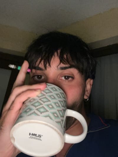 A dark-haired person is captured in a close-up view, angled slightly. Their eyes are prominent, focused towards the camera. They are drinking from a patterned mug labeled "HAUS Ceramics." A lit cigarette is held between their fingers. A blue jacket is visible in the frame. The background consists of muted tones. The overall impression is one of intimacy and casual lifestyle.