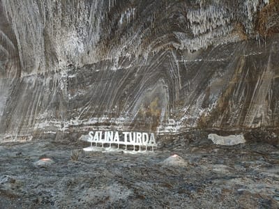 This media file depicts an indoor scene within a salt mine. The cavernous space is characterized by its rough, textured walls, which appear to be composed of layers of salt and rock, exhibiting a natural, organic pattern. Stalactite-like formations hang from the ceiling, some appearing crystalline and white, while others are darker and more mineralized. The floor is uneven and covered with debris, including small stones and salt crystals, suggesting an industrial or natural excavation site.
Prominently displayed in the center of the image is a white, three-dimensional sign that reads "SALINA TURDA." The letters are spaced out and supported by slender vertical structures. To the left of the sign, a small, reddish glow emanates from what appears to be a pile of salt or mineral. Another similar, smaller pile with a similar glow is visible further to the left. To the right of the sign, a white, intricately patterned object, possibly a decorative element or a piece of salt sculpture, is affixed to the wall. A thin, dark wire or pipe snakes across the lower portion of the scene.
The lighting is dim, contributing to the underground atmosphere, with the artificial light from the sign and the reddish glows providing the primary illumination. The overall impression is one of a vast, ancient, and geologically significant space, likely a tourist attraction given the prominent signage. The scene suggests a quiet moment, with no people visible, allowing the natural formations and man-made additions to be the focus. The location is identified as Turda, Romania, further confirming the context of a salt mine.