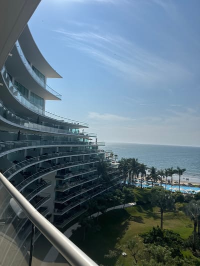 A high-angle shot shows a modern, curved building with glass-fronted balconies running along its facade. Beyond the building is a view of the ocean and a beach lined with palm trees. The beach transitions into a green lawn with more palm trees and some shade-providing trees. The sky is mostly clear with some wispy clouds. A portion of a railing is visible in the foreground.