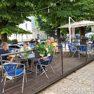 The image shows an outdoor cafe seating area under trees in Ruvo di Puglia, Italy. Four people are seated at tables on a wooden deck. A man with grey hair is seated on the left, facing a woman with short dark hair and a light-colored shirt. Further back, two men are seated at another table; one is wearing a yellow shirt and is facing away from the camera, while the other, facing the camera, is wearing a striped shirt and blue jeans. Blue chairs and tables are arranged on the deck, and a potted plant with white flowers sits on a table. In the background, buildings and parked cars are visible. The scene is lit by natural daylight, with strings of lights visible among the trees. A timestamp in the lower right corner reads "06.07.2026 09.52".