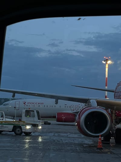 This is an image of a Rossiya Airlines aircraft parked on the tarmac at Yemelyanovo Airport in Russia. The aircraft is white with red accents and the airline's logo. The word "РОССИЯ" is clearly visible on the fuselage in red Cyrillic lettering. A tow tractor is attached to the front of the aircraft, suggesting it is being moved or prepared for departure. The sky is overcast and appears to be either early morning or late evening, indicating a twilight or dawn setting. The ground is wet, suggesting recent rain or damp conditions. The overall scene depicts an airport environment during a period of low light. The image appears to have been taken from inside a terminal or another vehicle, with a window frame visible in the foreground. The mood is neutral, focusing on the operational aspects of an airport.