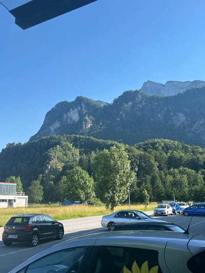 In Glanegg, Austria, a bright, clear day unfolds with a panoramic view of a majestic mountain range dominating the background under a cloudless blue sky. The lower slopes of the mountains are densely covered with lush green trees, gradually transitioning to rocky, exposed cliffs higher up. A prominent feature on one of the highest peaks is a small building, possibly a structure or observation post. In the foreground, a parking lot is visible, with several cars parked in neat rows. A black Volkswagen Polo with an Austrian license plate "S-448 WN" is parked on the left side of the lot. Other cars, including a silver sedan and various blue vehicles, are also present. Adjacent to the parking lot is a grassy field, with a modern building featuring a red and white striped awning on the far left. The scene captures a serene outdoor environment with a blend of natural beauty and human infrastructure.