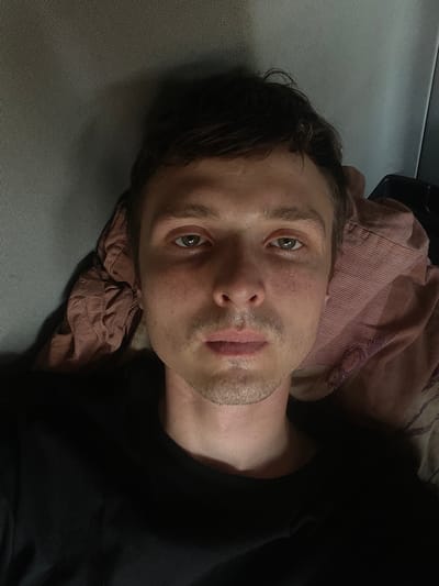 This is a close-up, sideways photograph of a person lying down. The subject is a young man with light skin, brown hair, and light green eyes. He has a few freckles on his face and stubble on his chin and jawline. He is wearing a black t-shirt. He appears to be lying on a textured gray surface, possibly a couch or bed, and is propped up by a pink and white striped fabric, which might be a pillow or blanket. The lighting suggests it is daytime, but the setting is indoors and appears dimly lit. The man's expression is neutral, and he is looking towards the camera. No text is visible in the image.