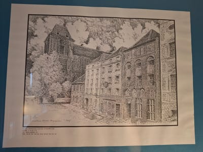 The image is a black and white ink drawing of an architectural scene. In the background, a large gothic-style building with a tall spire dominates the left side, partially obscured by trees. In the foreground and mid-ground, a row of multi-story buildings with brick facades and numerous windows lines a street. The style of the drawing is detailed, using numerous fine lines to create texture and shading.
At the bottom of the image, there is printed text which appears to be information about a hotel. It reads: "HOTEL GROMADA ZAJAZD STAROPOLSKI 87-100 TORUŃ UL. ŻEGLARSKA 10 TEL. 00-48 /56/ 260 60, FAX 00-48 /56/ 253 84". There is also a handwritten inscription above the printed text that appears to be a signature and a date, reading "~1998".
The prompt asks for the location in the city of Pila, Poland. However, the text on the image clearly states the hotel's location is in TORUŃ, not Pila. Therefore, based on the provided image, the location is Toruń, Poland.