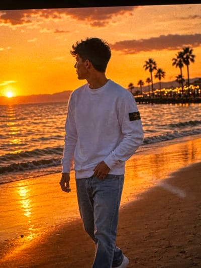 A young man stands on a sandy beach during sunset. He is facing away from the camera, looking out at the ocean. The sun is low on the horizon, casting a warm orange glow over the scene. The ocean is calm, with gentle waves lapping at the shore. Palm trees and lights from a boardwalk are visible in the background. The man is wearing a white sweatshirt with a patch on the left sleeve and blue jeans. The image does not depict any location in the city of Neubrandenburg, Germany; it appears to be a coastal scene, possibly in a tropical or Mediterranean region.