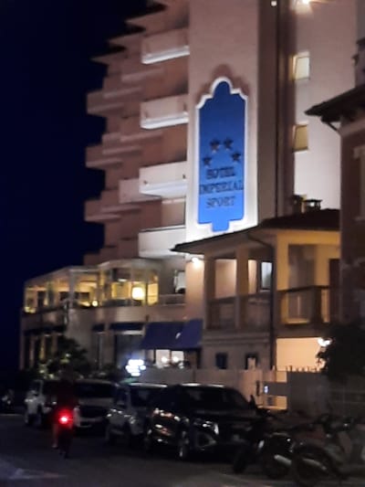 A nighttime street scene in Pesaro, Italy, features the facade of the "HOTEL IMPERIAL SPORT" with a distinctive blue sign and stars. The hotel has multiple stories with balconies and lit windows. In the foreground, several vehicles are parked or on the street, including a motorcycle with a red taillight, a white SUV, and other cars. The street is paved, and there are streetlights casting a glow on the scene. The overall impression is a quiet urban environment at night.