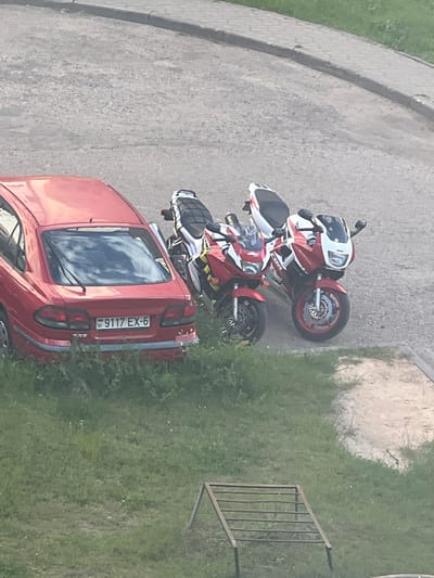 The image depicts a red car and two red and white motorcycles parked on an asphalt surface next to a patch of grass. The car, a hatchback, is parked closer to the grass, with its rear facing the viewer. The license plate of the car is visible and reads "9117 EX-6". The two motorcycles are parked behind the car, side by side, and appear to be sports bikes. One motorcycle is slightly in front of the other, and both are oriented similarly to the car. The scene is outdoors, with a paved area on the left that transitions into the asphalt where the vehicles are parked. The right side of the image is dominated by green grass and some foliage. In the background, there is a hint of a fence or railing structure. The lighting suggests it is daytime, likely with bright sunlight, as indicated by the reflections on the car. The overall setting appears to be a residential area or a parking lot in a town. There are no people visible in the image. The image is taken from an elevated perspective, looking down on the vehicles and the surrounding environment. The weather appears clear and pleasant, typical for a sunny day. The location context provided is Asipovichy, Belarus.