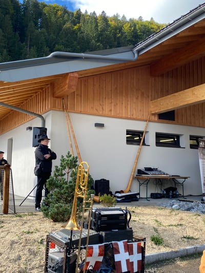 This image depicts an outdoor event setup against a modern building in Abondance, France. The building features a grey tiled roof with a prominent gutter, an overhanging eave with a visible wooden underside, and a wall partially clad with vertical light-brown wooden planks and partially with plain white stucco. Two rectangular dark-framed windows are visible in the white stucco section of the wall, along with two black wall-mounted light fixtures.
In the background, a dense green forest covers a steep hillside under a blue sky with scattered clouds. The ground in the foreground and mid-ground is covered with light-colored gravel.
Musical equipment is prominent. Three long, light-brown wooden alphorns lean against the building: two on the left side, angled towards the wooden cladding, and one on the right side, leaning against the white stucco. In the foreground, slightly left of center, a golden brass trombone stands on a metallic rack holding a sound mixer. Below the trombone, a black accordion rests on a box. A banner, consisting of several small white flags with red crosses, is draped across the front of this equipment stand. Further right, a collapsible wooden table supports a larger mixing console and other audio equipment, with cables visible on the ground. A black speaker is positioned on the ground to the right of this table. Another black speaker is mounted on a stand to the left of the primary human subject.
One main male subject is visible in the mid-ground, slightly left of center. He is wearing a black hat, glasses, a black jacket, and dark trousers, with a dark shoulder bag. He stands with his arms crossed, looking towards the right. Part of a second male subject is visible on the far left, wearing a dark beret and a dark jacket with a gold emblem, standing behind a wooden post.
A small, conical evergreen bush is situated between the main male subject and the foreground musical equipment. On the far right, a narrow vertical banner with "1425 LES CROSES" is affixed to the building's wall.