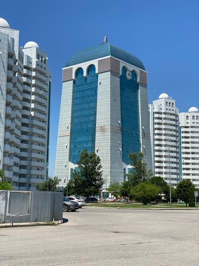 This is a vertical, eye-level photograph taken outdoors on a sunny day, likely in the afternoon due to the clear blue sky and strong shadows. The scene features a modern cityscape dominated by tall, white apartment buildings and a prominent glass-fronted office building with a large clock face near its top. The apartment buildings have many balconies, giving them a layered appearance. The office building has a curved glass facade in shades of blue and grey, with distinct architectural features like arched window openings.

In the foreground, a paved road runs vertically along the right side of the image, with a sidewalk or grassy area separating it from the buildings. A white car is parked on the side of the road, casting a shadow. Some of the windows on the office building are open. To the right of the office building, mature green trees provide a natural element. A temporary metal fence is visible in the middle ground.

There are no people visible in the photograph, and the overall mood is one of urban development and quiet activity. The location is indicated as Shali, Russia, which provides a geographical context for the modern architecture. The time of day is suggested by the bright sunlight and sharp shadows. The weather appears to be clear and warm. There is no visible text in the image.