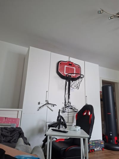The image depicts a room interior, likely a home in Helmstadt, Germany, given the prompt's location reference. The scene contains a variety of objects: a red and black gaming chair with a white desk and controller on top of it; a black cylindrical object, possibly a punching bag or workout equipment; a miniature basketball hoop in red and black; several clothes hangers; a box containing toy cars; clothing items (specifically a grey knitted blanket and socks); and white storage cabinets. The room is illuminated by a ceiling light fixture with multiple spotlights. There is no visible indication of any people actively participating in any action. The image appears to be a snapshot of a storage or organization space, suggesting a living area or hobby room.