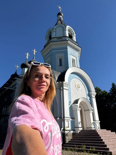 A woman is taking a selfie with a church in the background in St. Ozerki, Russia. She is wearing a pink t-shirt and white sunglasses pushed up on her head. The church has blue and white accents and a bell tower with a gold cross on top. The sky is clear and blue.