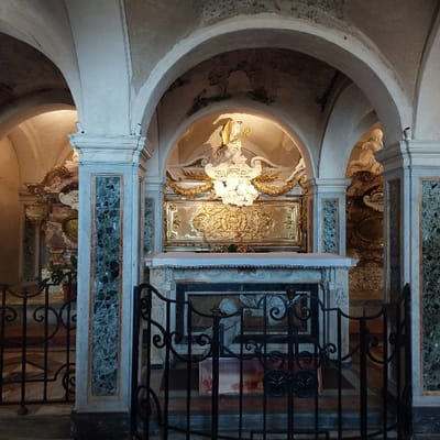 The image depicts an interior view of the Crypt of the Cattedrale di San Ciriaco in Ancona, Italy. The scene features three distinct, arched alcoves, each housing an ornate tomb or altar, separated by large, fluted columns composed of white marble with prominent dark green veining.

In the central alcove, a white marble altar-like structure is positioned behind a black wrought iron gate. Above this altar, a rectangular golden panel with intricate relief carvings is surmounted by a white marble sculpture of a winged figure, possibly an angel, holding an object, with a scroll inscribed "SANCTE CUPITI ET FRATRES MARTYRES" below. Gilded laurel wreaths flank the sculpture. A small red object, resembling a flower, rests on the altar. The wrought iron gate, with its decorative scrollwork and vertical bars, spans the lower foreground, partially obscuring the base of the central altar, which appears to feature an embedded relief or inscription.

To the left, beyond a marble column, a second alcove is partially visible, containing similar ornate gold and marble elements and another section of the wrought iron gate. A small potted plant is present within this section. To the right, a third alcove is also partially visible, showcasing another white marble sculpture of a reclining figure, similarly adorned with gold detailing.

The vaulted ceiling is a pale, textured material, exhibiting signs of age. The lighting is low, with strategic illumination highlighting the altars and sculptures. The floor appears to be made of dark stone or tiles. No individuals are present in the scene.