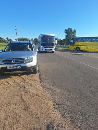 A silver Renault Duster SUV is parked on the side of a paved road, its front facing right. The SUV has a license plate reading "8039 PH-7" and a driver, a man with light skin and short dark hair, is visible behind the wheel. To the right of the SUV, a white bus with the number "702" and Cyrillic text "ЛІДА-СЛОНІМ" is driving on the road. Further to the right, a yellow bus is also driving on the road. The road is lined with trees and vegetation. The sky is clear and blue. The ground beside the road where the SUV is parked is dirt and shows tire tracks. This scene is located in Цімкавіцкі сельскі Савет, Belarus.