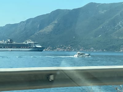 A long cruise ship, identified by the text "CELEBRITY CONSTELLATION" on its side, is docked or moving slowly in a bay, with its port side visible. The ship has multiple decks and is predominantly white with dark blue accents. To the right of the cruise ship, a smaller white motor yacht is speeding through the water, leaving a wake. In the background, a verdant, mountainous landscape rises steeply from the coastline, dotted with houses and buildings characteristic of a Mediterranean town. The water is a clear blue, and the sky is a pale blue, suggesting a sunny day. A blurred metallic barrier is in the foreground, obscuring the lower part of the view. The scene is consistent with the Bay of Kotor near Donji Orahovac, Montenegro, known for its dramatic coastal scenery and large vessels.