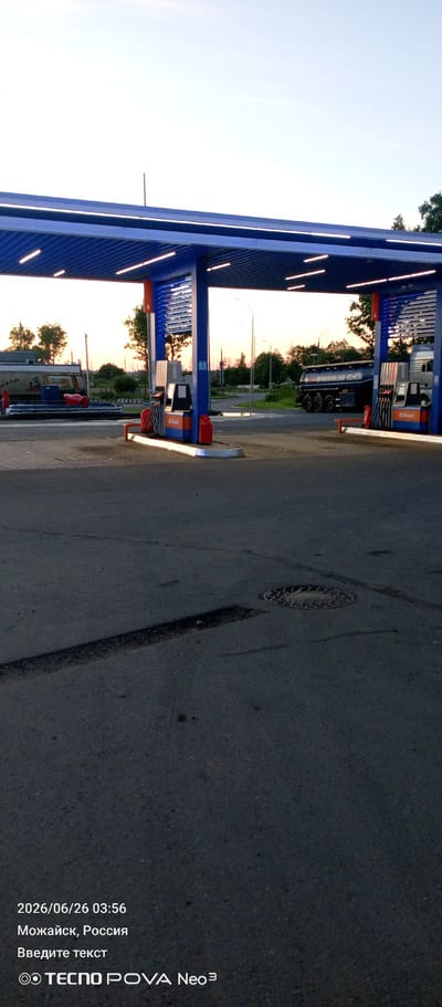 A dusk or dawn view of a gas station in Mozhaysk, Russia shows a bright blue canopy over multiple gas pumps. The sky is a pale blue, transitioning to a lighter hue near the horizon where a faint orange glow suggests the sun is either rising or setting. Trees and some structures are visible in the background, silhouetted against the sky. Two trucks are present; one, a tanker truck with Cyrillic lettering that appears to say "ОГНЕОПАСНО" (Flammable), is parked near the pumps, and another vehicle, partially visible, is further back on the right. The ground is paved asphalt, with some tire marks and a manhole cover visible in the foreground. On the bottom left of the image, the date "2026/06/26 03:56", the location "Mozhaysk, Russia", and the text "Введите текст" (Enter text) are superimposed, along with a logo that reads "© TECNO POVA Neo³".