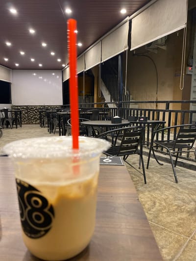 Here is a description of the image:
The image shows a close-up of a plastic cup filled with a light-brown beverage, presumably an iced coffee, sitting on a wooden table. A red straw is inserted into the drink. The cup has a black logo on its side with a circular pattern resembling eyes. The background reveals an outdoor cafe or patio setting. There are several black metal chairs and tables arranged in rows, suggesting a seating area. A set of metal stairs is visible in the background, leading to a higher level. The ground appears to be paved with tiles. On the left side of the frame, a building wall is visible with multiple small lights affixed to it. The location appears to be Keudeublang, Indonesia, based on the "MEUGANG Keudeublang" sign visible on the table. No people are visible in the frame.