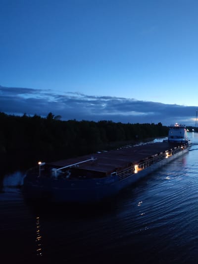 A long, dark barge is moving through a waterway at dusk, likely a canal or wide river. The barge, loaded with a dark cargo, is positioned diagonally in the frame. Small lights illuminate its sides. Dense, dark trees line the banks of the waterway in the background. The sky is a gradient of deep blues, with darker clouds on the horizon. The water surface reflects the lights of the barge and the sky. The location is potentially Annensky Most, Russia, based on the waterway.