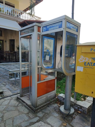 This image depicts an outdoor scene in Litochoro, Greece, featuring two cardphone booths and a yellow Hellenic Post mailbox. The setting appears to be a paved area with stone tiles, suggesting a public space like a street or plaza. Behind the booths, a building with a white railing and a tiled facade is visible, indicating a residential or commercial area.
The foreground is dominated by two phone booths. The one on the left is open, revealing a silver metal frame and orange panels. The booth on the right is mostly enclosed, with a glass front showing a black telephone handset and a keypad. A woman is faintly visible through the glass, reflected in the phone booth. Both booths have signage that reads "ΚΑΡΤΟΤΗΛΕΦΩΝΟ" and "CARDPHONE" in blue text.
Adjacent to the right phone booth is a bright yellow mailbox, also bearing Hellenic Post branding. Graffiti in white spray paint is visible on the mailbox. The ground is scattered with dry leaves, and green grass is present behind the mailbox. The lighting suggests it is daytime, with sunlight casting shadows and highlighting the objects. The overall atmosphere is that of a quiet, everyday street scene in a Greek town.