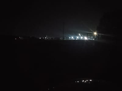 The image depicts a nighttime scene in Ertis, Kazakhstan, characterized by darkness and artificial lighting. A flat, dark foreground suggests a ground surface. In the middle ground, a line of illuminated structures and streetlights are visible against the night sky, indicating a populated or industrial area. The lights are bright and some create starburst effects, suggesting they are high-intensity lamps. The background is predominantly black, with faint hints of clouds or atmospheric haze. A dark silhouette of foliage, possibly a tree, is present on the right side of the image. The overall impression is of a dimly lit urban or industrial landscape at night.