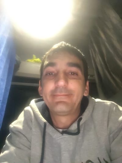 The image displays an adult male, captured from the chest up, centrally framed and directly facing the camera. He has short, dark hair and dark eyes, exhibiting a slight smile. He is dressed in a light grey hooded sweatshirt with a black drawstring and a visible dark collar beneath. The background suggests an enclosed interior space, consistent with a vehicle cabin. To the subject's left, a blue fabric panel or curtain is present. To his right, a darker, pleated fabric, possibly a curtain or bunk lining, is visible. An intense overhead light source in the upper center creates significant glare and lens flare. A light-colored shelf or bunk is partially visible behind the subject's head, containing a small, crumpled yellow object. The image contains no visual elements to confirm the specified location of Überherrn, Germany.