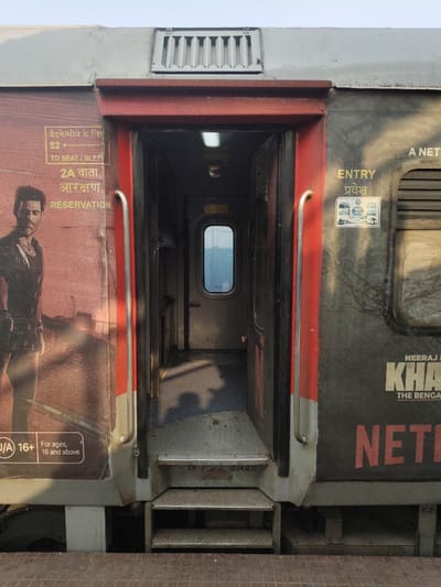 An external view of an Indian Railways train car is captured at a platform in Bardhaman, India. The central focus is an open passenger door, framed by a prominent red border, revealing a dimly lit interior corridor culminating in a small, oval-shaped window. Two corrugated metal steps descend from the train car to a reddish-brown textured platform.
The train's exterior is divided into two distinct advertising panels:
On the left, a reddish-brown panel features a large image of an Indian actor (Hrithik Roshan, associated with the movie "Vikram Vedha") with text indicating "2A VATA आरक्षरण" (2A RESERVATION) and "TO SEAT / SLEEP," alongside an "For ages, 16 and above" advisory.
On the right, a dark grey panel displays "ENTRY" in English and Hindi ("प्रवेश"), a small multi-image sticker, partially obscured text including "NEERAJ KHAN," "THE BENGAL," and a large red "NETFLIX" logo.
Above the door, a grey ventilation grille is visible on the roofline. Markings "ER" and "-224187C" are present near the steps, likely identifying the Eastern Railway and a coach number segment. No individuals are actively present, though shadows are cast across the steps and into the train's interior by an unseen light source. The sky above the train is light blue.