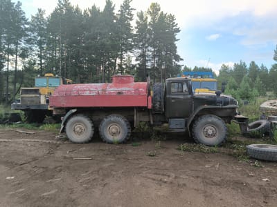 Two large, older model military trucks are parked on a dirt lot with pine trees in the background. The truck in the foreground is dark green with a large red cylindrical tank mounted on its chassis. It has six large, knobby tires and appears to be a fuel or water truck. The truck behind it is yellow and appears to be a loader or construction vehicle. The sky is partly cloudy. The location is identified as Gremiuchiy, Russia.