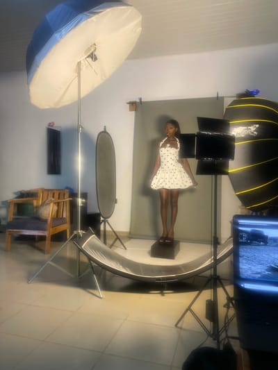 A young Black woman stands on a dark platform in a photography studio, posing for a picture. She is wearing a short, white dress with black polka dots and black sandals. The studio is set up with various lighting equipment, including a large softbox with yellow accents, a ring light, and a reflector. A gray backdrop is behind the woman, and a monitor displaying an image is visible in the background. Wooden chairs are arranged to the side. The floor is tiled. The lighting suggests an indoor setting, likely during the day. There is no visible text in the image. The woman appears confident and poised. The overall impression is a professional studio photoshoot.