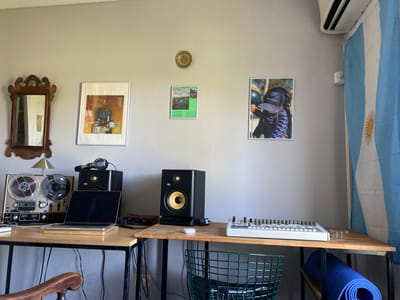 A home music production studio is set up against a gray wall adorned with a mirror, three framed pictures, and a decorative plate. The pictures feature different subjects, one of which seems to be a photo of a child wearing a Peugeot cap. A studio monitor, vintage tape machine, laptop, and headphones sit on a wooden desk. A green wire chair and a rolled-up blue yoga mat are visible under the desk. An Argentine flag hangs on the right side of the wall, suggesting the studio is located in Argentina. While the location Boulogne Sur Mer is mentioned, there is no visual evidence in the image to confirm this fact.