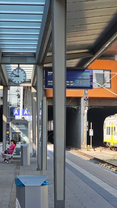 A train station platform in Neu-Ulm, Germany features a large circular clock with Roman numerals showing approximately 9:10. A digital display board shows departure information: "08:54 RS 71", "09:20 Ulm Hbf", and "09:55 Ulm Hbf". To the left of the digital display, a vending machine labeled "SNACKS & DRINKS" is visible, along with a seating area. A young woman with dark hair, wearing a pink shirt and black shorts, is sitting on a bench facing a vending machine, looking at her phone. An outdoor trash receptacle with a blue liner is positioned in the foreground. In the background, a train track leads into a dark tunnel. A yellow and white train car is partially visible at the edge of the platform. The platform is covered by a modern, slatted roof structure supported by metal pillars. The overall environment appears to be an outdoor public transportation hub under bright daylight.