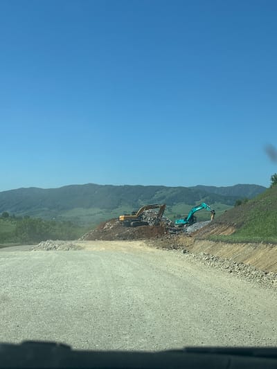 The image depicts a construction site in the mountainous terrain of Sarasa, Russia. Two excavators are actively engaged in demolition and excavation work, with piles of rubble and earth surrounding them. A light-colored gravel road leads into the foreground, suggesting ongoing infrastructure development. The background features rolling green hills and a clear blue sky, indicative of a sunny day. The scene is devoid of human presence, with the focus solely on the heavy machinery and the landscape.