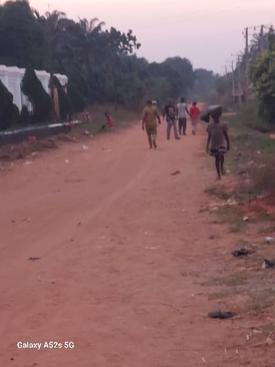 The image depicts an unpaved, reddish-brown dirt road in Umudioka, Nigeria, under a muted, light-colored sky suggesting either dusk or dawn.
Seven individuals are present. In the midground on the left side of the road, a person in a red shirt is seated or crouched near a low white concrete barrier. Behind them, a white structure with multiple triangular architectural elements is partially visible, largely obscured by dense green foliage.
On the right side of the road, in the midground, a young individual wearing dark clothing walks towards the viewer, carrying a dark, possibly textile-wrapped object on their head. The road's edge on this side contains sparse grass and scattered dark debris. Further in the background on the right, utility poles with overhead wires are visible among more green vegetation.
In the center of the road, a group of five individuals walks away from the viewer. The foremost figure on the left wears a light brown or khaki uniform-like outfit. The other four individuals wear a mix of dark and light shirts, including one red and one white, with light-colored pants. The foreground of the road consists of reddish-brown dirt with minimal scattered debris. The watermark "Galaxy A52s 5G" is visible in the lower-left corner.