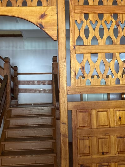 This image depicts an interior architectural scene in Bayronovka, Russia. On the left, a wooden staircase ascends, featuring worn, light-colored treads and risers with visible peeling finish. A wooden balustrade, consisting of square-topped posts and three horizontal planks, borders the right side of the staircase. Above the staircase, a wooden archway with decorative scalloped and rounded cutouts along its top edge is visible against a plain white wall. The right side of the frame is occupied by a large, two-tiered wooden partition or screen. Its upper section features an ornate lattice design with interconnected oval and teardrop-shaped cutouts. The lower section comprises multiple raised rectangular panels. All wooden elements exhibit light to medium brown tones, displaying prominent grain patterns and knots. The background throughout the scene consists of light-colored walls. No individuals are present.