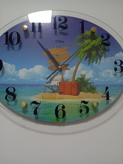 This is a close-up, eye-level, medium shot of a round wall clock with a beach scene depicted on its face. The clock features large, black Arabic numerals indicating the hours. The background of the clock face shows a tropical island with a palm tree, a beach chair, a suitcase, and a wooden signpost, set against a blue sky with clouds and a turquoise ocean. The clock hands are visible, with the hour hand pointing roughly between the 11 and 12, and the minute hand pointing towards the 10. The clock is mounted on a white wall, casting a subtle shadow. The location of this clock in Toktogul, Kyrgyzstan is not determinable from the image alone.