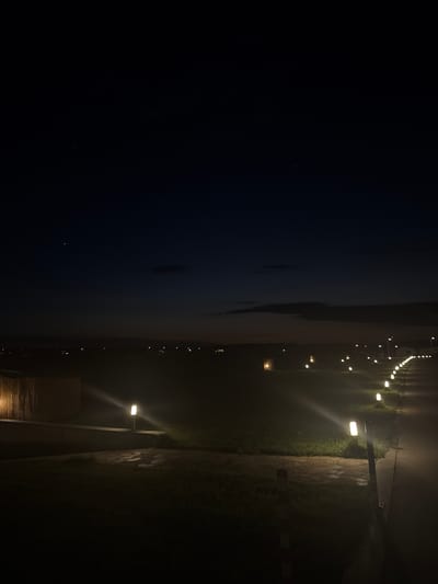 The image captures a dark, atmospheric scene in Topola, Bulgaria. The foreground is dominated by a pathway illuminated by a series of modern, rectangular lamps, spaced evenly along its right side. These lamps cast a warm, white light onto the asphalt path and the surrounding grass, creating a sense of depth and leading the eye into the distance. To the left of the path, there appears to be a low, textured wall or structure, also softly lit.
Beyond the immediate path, a dimly lit field stretches out, with scattered lights suggesting a distant village or settlement. The sky above is a deep, dark blue, transitioning to a lighter hue near the horizon, indicative of dusk or early night. A few faint clouds are visible against the dark sky. The overall impression is one of tranquility and a slightly ethereal quality due to the interplay of artificial light and the natural darkness of the evening. No people or animals are visible in the frame. The environment appears to be an outdoor space, possibly a park, a resort, or a well-lit residential area.