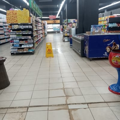 The scene depicts an interior view of a supermarket aisle located in Kisa Central ward, Kenya. The floor is covered with light-colored square tiles, exhibiting visible discoloration and moisture in several areas, particularly in the foreground and mid-ground. A yellow A-frame caution sign, marked "KANGUKA" and featuring a pictogram of a falling person, is centrally positioned on the aisle floor. To the left, a long shelving unit with multiple tiers of black metal shelves is fully stocked with various packaged products, including numerous yellow-boxed items on the top shelf. To the right, a grey, open-top commercial refrigeration unit is situated alongside a blue "Cadbury Ice Cream" branded freezer unit, which displays "Ice Cream" text and product imagery. In the bottom right foreground, a portion of a red and blue, goblet-shaped display stand labeled "Big Giant" is visible. A dark brown, possibly woven, cylindrical container is partially visible in the bottom left foreground. Further into the background, additional aisles, refrigeration units, a silver cylindrical bin, and a blue Red Bull branded cooler are discernible. The area is illuminated by overhead fluorescent lighting. No individuals are present in the image.