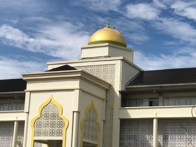 A mosque, likely in the city of Reuleut Timu, Indonesia, is captured from a low angle under a partially cloudy blue sky. The building features a prominent golden dome adorned with a green crescent moon. The facade is cream-colored with intricate white latticework and decorative yellow arches. Dark, sloped roofs are visible, as are rows of windows on the upper levels. The overall impression is one of modern Islamic architecture against a bright, natural backdrop.