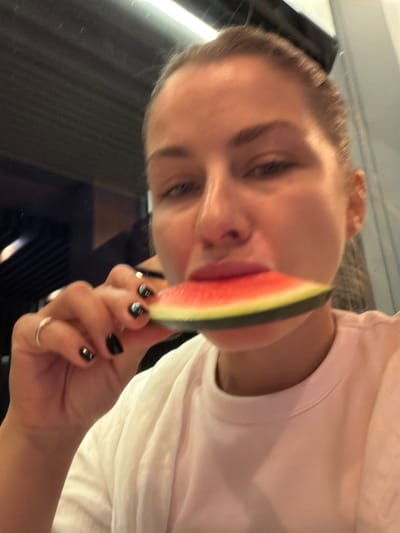 A woman is shown from the chest up, holding a slice of watermelon to her mouth. She is wearing a white t-shirt and has black nail polish and two rings on her left hand. The background is blurred, but appears to be indoors, possibly a restaurant or cafe. There is a light fixture visible at the top of the image. The scene is not identifiable as being in Žunovnica, Bosnia and Herzegovina.