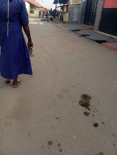 In Gwagwa, Nigeria, a street scene unfolds with a person in the foreground walking away from the camera, their blue and red patterned dress dominating the left side of the frame. The individual is wearing brown sandals. Behind them, other people are visible further down the dusty street, some tending to stalls or engaged in conversation. Buildings line the sides of the street, some with corrugated metal roofs and others with more permanent structures. A drainage channel is visible along the right side of the street, partially filled with water and debris. Several dark, wet patches, likely oil or liquid spills, are present on the asphalt surface in the middle and lower right portions of the image. The overall environment appears to be an open-air market or commercial area.
