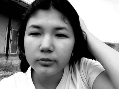 A black and white, close-up, eye-level selfie of a young person with dark hair is captured outdoors. The person's face is centered, with their right hand resting on the back of their head. They are wearing a light-colored t-shirt. The background features an older building with a door and window, some overgrown vegetation, and a gently sloping hill. The sky is bright and overcast. The setting appears to be in Mady, Kyrgyzstan.