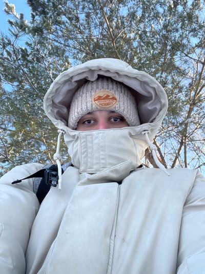 A close-up selfie shows an individual dressed in heavy winter attire in Novochuvashskii, Russia. The person is wearing a cream-colored puffer jacket with the hood pulled tightly around their face, exposing only their eyes. Beneath the hood, a light beige knitted beanie is visible, featuring a circular patch with a mountain graphic and the text "MOUNTAIN PATHWAYS". A grey backpack strap with a black buckle is visible over the individual's left shoulder. The background consists of snow-dusted pine trees and bare deciduous tree branches against a clear blue sky, indicative of a cold, outdoor winter environment.