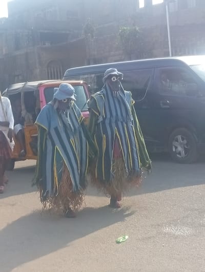 A daytime street scene in Itakpe Junction, Nigeria, depicts two individuals in elaborate masquerade costumes walking forward on a paved road. Each costumed figure wears a light blue brimmed hat featuring white and yellow concentric circles simulating eyes, a vertically striped tunic in dark blue, light blue, and yellow, and a lower garment of natural fibers (possibly raffia or straw) extending to their ankles. Dark reddish-brown sandals are visible on their feet. Their faces are obscured. The figure on the left is slightly ahead of the figure on the right.

To the left, a portion of a pink and yellow three-wheeled auto-rickshaw (keke napep) is visible, with at least one passenger discernible inside. Further left, partially obscured, an individual in a white shirt and dark lower garment walks. To the right, a dark blue passenger van is either stationary or moving slowly behind the costumed figures, with a faint human profile visible through its front passenger window.

The road surface is light grey/tan asphalt, with a crumpled green object near the bottom right. Long shadows are cast from the figures and vehicles towards the foreground, indicating bright sunlight. The background features multi-story buildings with weathered facades, sparse vegetation, and a vertical pole on the far right.