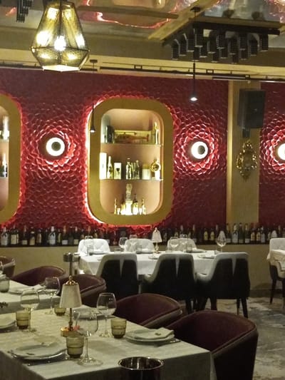 The image captures an upscale restaurant or dining area, likely in the evening or at night, given the artificial lighting. The dominant feature of the interior is a striking red wall, highly textured with a repetitive, circular, almost petal-like pattern. Embedded within this red wall are several gold-rimmed, vertically elongated oval alcoves, acting as illuminated display shelves. The central alcove visible in the main image is well-lit and filled with various bottles, glasses, and decorative items, suggesting a bar or a display of spirits.
Between these illuminated alcoves, and along the bottom portion of the red wall, a long shelf displays numerous wine or spirit bottles arranged in a neat row. Above these bottles, the red textured pattern continues, featuring circular light fixtures that emit a soft, warm glow, enhancing the ambiance.
The ceiling above is reflective, possibly mirrored or with a highly polished surface, showing reflections of the room below. Several light fixtures hang from the ceiling; a prominent, geometric, cage-like pendant light with multiple bulbs is visible in the upper left. Another, more modern, multi-directional light fixture with a rectangular base and numerous small spotlights is visible towards the right side of the ceiling. A dark, possibly square or rectangular speaker is mounted high on the wall to the right, adjacent to a decorative gold ornament.
In the foreground and midground, several dining tables are set. They are covered with crisp white or light-colored tablecloths, suggesting elegance. Each table is meticulously set with white plates, various types of glassware (wine glasses, water glasses, tumblers), and cutlery. Small, elegant table lamps with textured shades provide additional soft lighting on some of the tables. The chairs are upholstered, featuring a combination of dark and lighter colors, with a curved back design, providing a comfortable yet sophisticated look. Some chairs appear to be a dark gray or black, while others are a deep maroon or burgundy, contrasting with the light table settings. The floor is not clearly visible but appears to be a neutral, possibly tiled or polished concrete surface. There are no people visible in the scene.