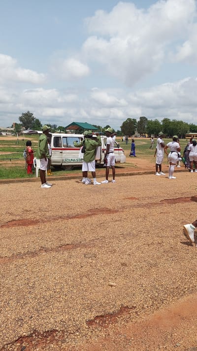 In Unguwar Shanu, Nigeria, on a bright, partly cloudy day, a group of individuals, predominantly young adults in uniform, are gathered outdoors on a rough, gravelly surface. Several individuals are wearing green shirts, white shorts, and green caps, suggesting they are part of a uniformed service or organization, possibly national service members. Some of these individuals are standing near a white van with the partial text "KADUNA STAT" visible on its side. The van appears to be parked on a paved area or road. In the background, there are trees, buildings with green roofs, and other individuals in casual attire. The overall scene suggests a gathering, possibly a briefing or waiting period, in an open, semi-urban environment.
