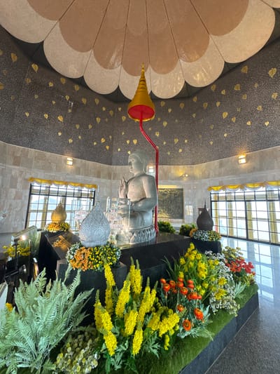 The image depicts the interior of a Buddhist shrine in the city of Chang Khoeng, Thailand. Dominating the center is a large, stone statue of a seated Buddha. The statue is positioned on a black altar adorned with a profusion of colorful artificial flowers, primarily in shades of yellow, orange, and white, interspersed with green ferns. Behind the altar, a tall, ornate, golden spire with a red base is suspended by a red rod, positioned directly above the Buddha's head.
The shrine's walls are clad in light-colored marble tiles. The ceiling features a distinctive petal-like design, with alternating sections of cream and beige. The dark gray ceiling above is decorated with numerous small, gold leaf-shaped motifs. Large windows with a grid pattern are visible on the sides, allowing natural light to enter the space and offering a view of the outdoors, suggesting a mountainous or hilly terrain beyond. Several decorative items, including a golden stupa-like ornament and a dark, finial-topped urn, are placed on the altar. A large, carved relief depicting what appears to be a Buddhist narrative is mounted on the wall in the background. The floor is reflective, indicating a polished surface, possibly granite or marble. No people are visible in the image. The scene conveys a sense of reverence and peacefulness.