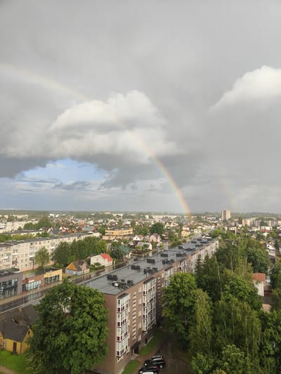 The image shows an aerial view of a city with a double rainbow in a cloudy sky. The city appears to be a mix of residential buildings, including apartment blocks and smaller houses, with trees and greenery interspersed. The streets are wet, suggesting recent rain. In the foreground, there is a large apartment building with a dark roof. There are a few cars parked on the street. The distant part of the city shows more houses and buildings, with a dense tree line in the background. The main rainbow is prominent, arching across the sky, with a fainter secondary rainbow visible to the right. The sky is dominated by grey clouds, with some patches of lighter sky. The lighting suggests it is daytime, possibly late afternoon or early evening, given the dramatic sky. Based on the architecture and urban planning, this scene is characteristic of residential areas in Panevėžys, Lithuania, particularly areas with Soviet-era apartment blocks and surrounding suburban housing.