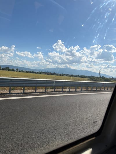 The image depicts a view from inside a vehicle, looking out onto a roadside scene. The foreground is dominated by the asphalt of a road with a white line marking its edge. A silver metal guardrail runs parallel to the road, separating it from a grassy embankment. Beyond the embankment, a field of light green crops stretches towards a hazy, mountainous horizon. The sky is a bright blue, dotted with fluffy white cumulus clouds. Sunlight streams down, creating some lens flare and highlights on the windshield. A power line and utility pole are visible in the distance. The scene is likely located near Yana, Bulgaria, given the context. There are no people visible in the image.